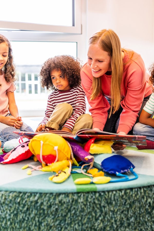 kita-kinderzimmer-hamburg-brand-shooting-kinder-erzieher Eine Pädagogin liest einer Gruppe ein Buch vor in der Kita kinderzimmer Hamburg