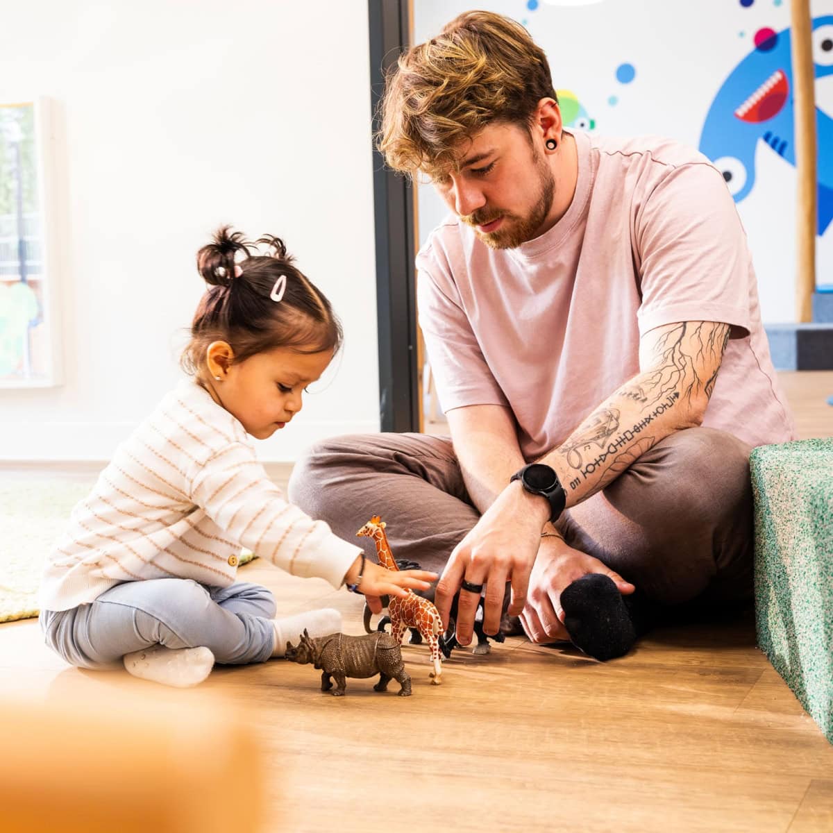 kita-kinderzimmer-hamburg-brand-kjell-kind-spielen Pädagoge spielt, sitzend auf dem Boden, mit Kleinkind und Spielzeugtieren